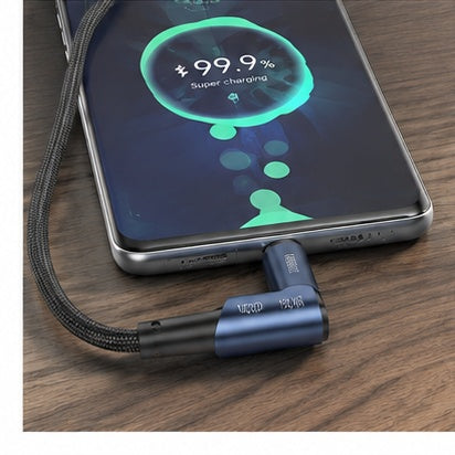 A close-up of a smartphone charging on a wooden surface, connected to a black braided cable with a dark blue angled connector. The phone screen shows a green '99.