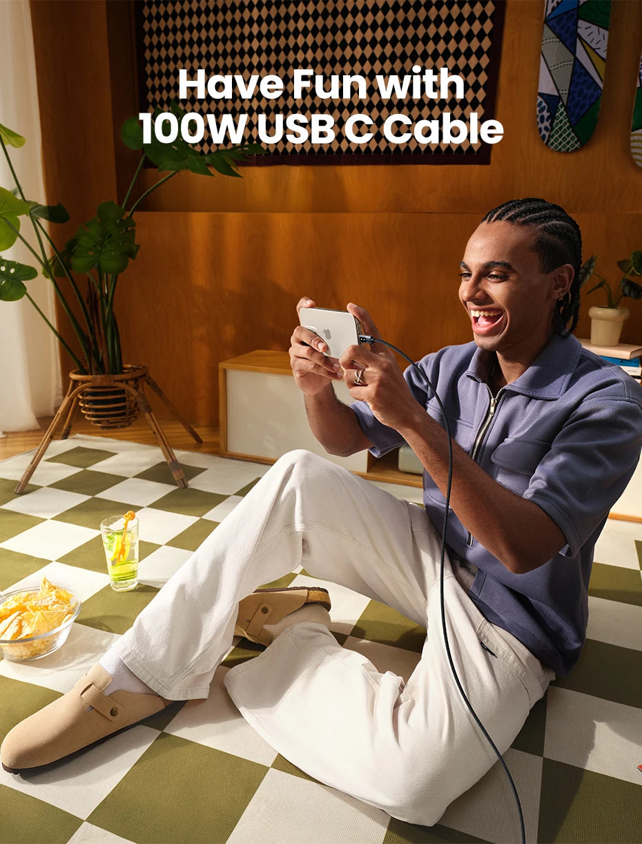A man sitting on a green and white checkered floor is smiling while holding a white smartphone and connecting a black cable, with the text 'Have Fun with 100W USB C Cable' overlaid at the top. He is wearing a blue shirt and white pants in a cozy indoor setting with plants and snacks.