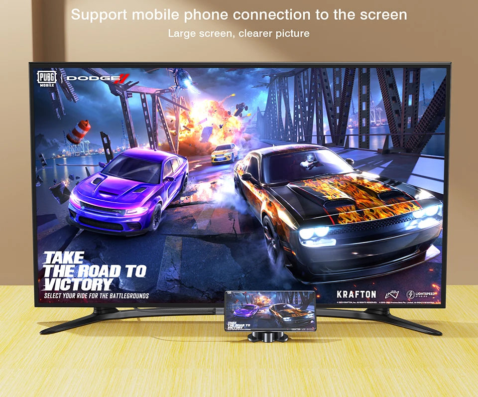 A large flat-screen TV on a light wood surface against a beige wall, showing a racing game scene with two cars and on-screen PUBG and Dodge branding. A small phone is placed in front of the TV to demonstrate mobile connection.