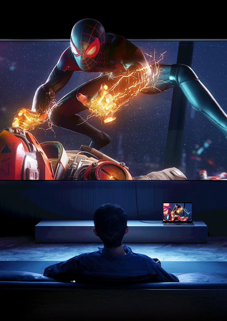 A person sits in a dark room facing a large wall-mounted screen showing an action scene of Spider-Man with energy effects, while a smaller laptop on a stand shows the same content.