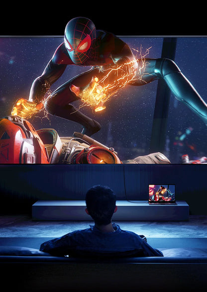 A person sits in a dark room facing a large wall-mounted screen showing an action scene of Spider-Man with energy effects, while a smaller laptop on a stand shows the same content.
