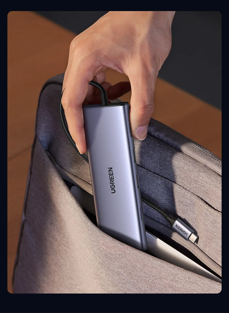 A hand inserting a slim silver UGREEN power bank into a gray laptop backpack, shown in a close-up shot with a dark border.