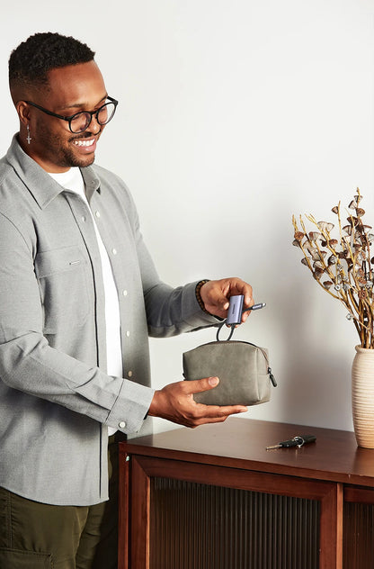 Man holding a small gray bag next to a wooden desk with a vase of dried flowers.