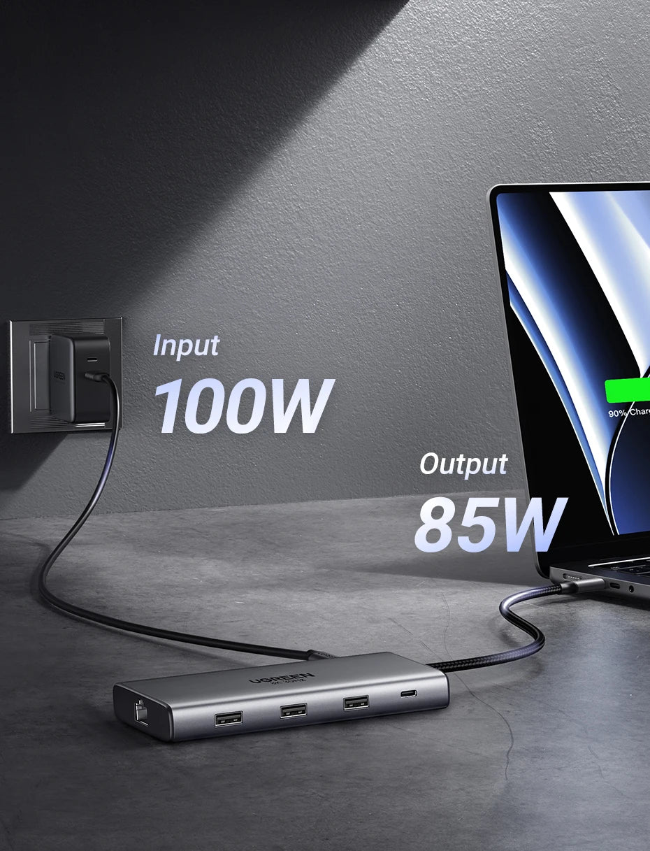A 100W to 85W laptop power adapter hub with multiple USB-C ports, shown connected to a wall outlet and a laptop on a dark gray surface with text overlays indicating power input and output.