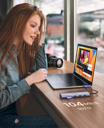 A woman sits at a wooden desk using a laptop, with a portable external SSD drive placed on the desk in front of her. The drive is silver and black, shown connected to the laptop with a speed overlay reading 104MB/S SSD/TF.